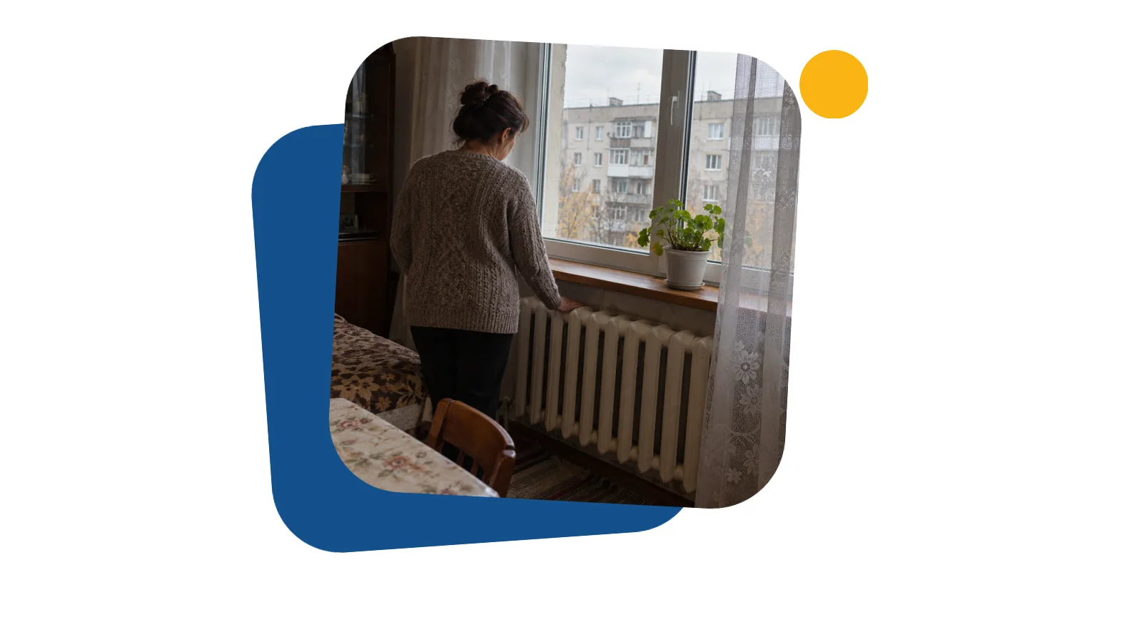 Жінка зі спини стоїть біля батареї під вікном у кімнаті орендованої квартири в багатоповерхівці і торкається її долонею