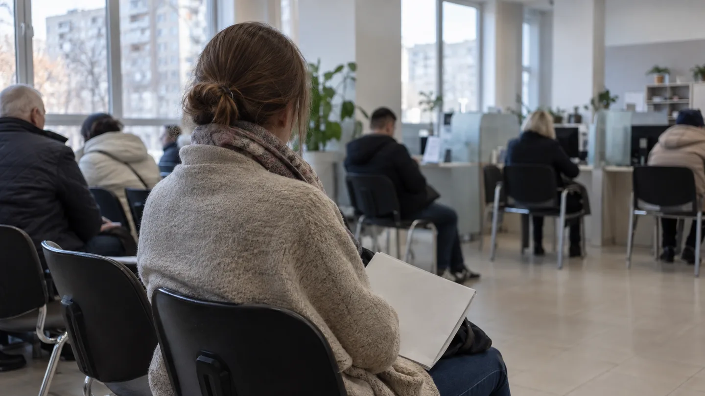 Жінка зі спини сидить у черзі в залі ЦНАП з папкою документів на колінах