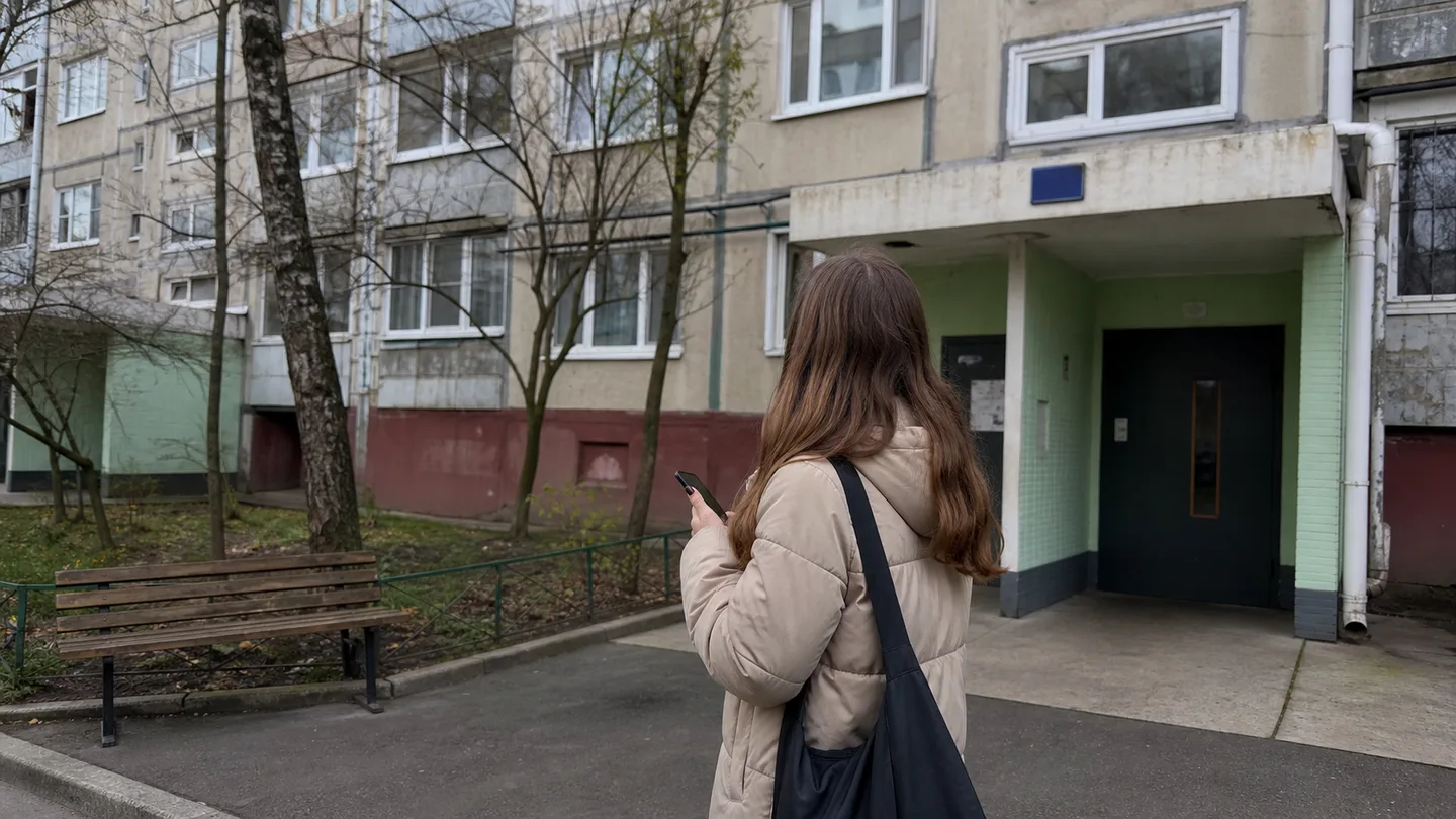 Жінка з телефоном стоїть перед під'їздом багатоповерхівки й звіряє номер будинку