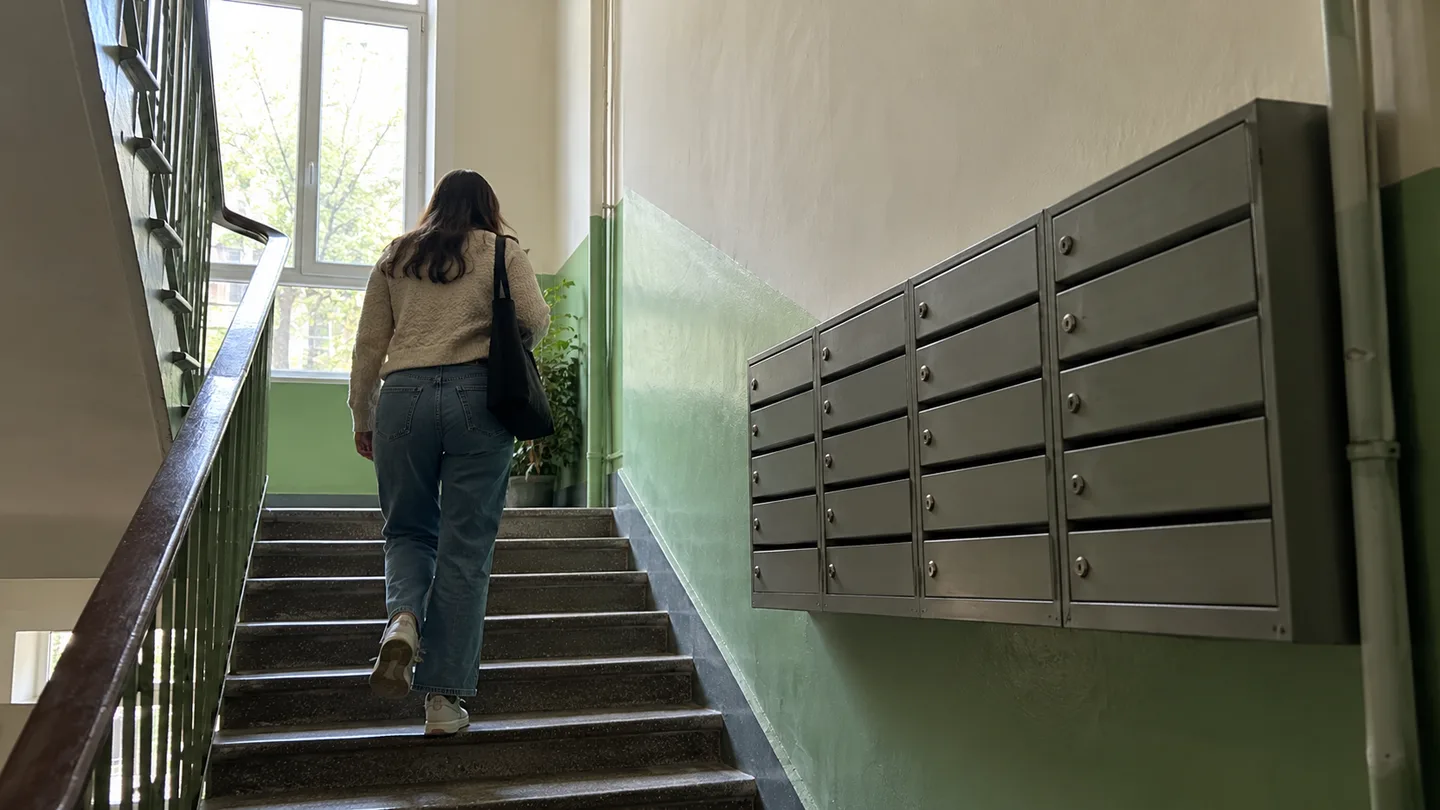 Жінка зі спини підіймається сходами світлого під'їзду з поштовими скриньками вздовж стіни
